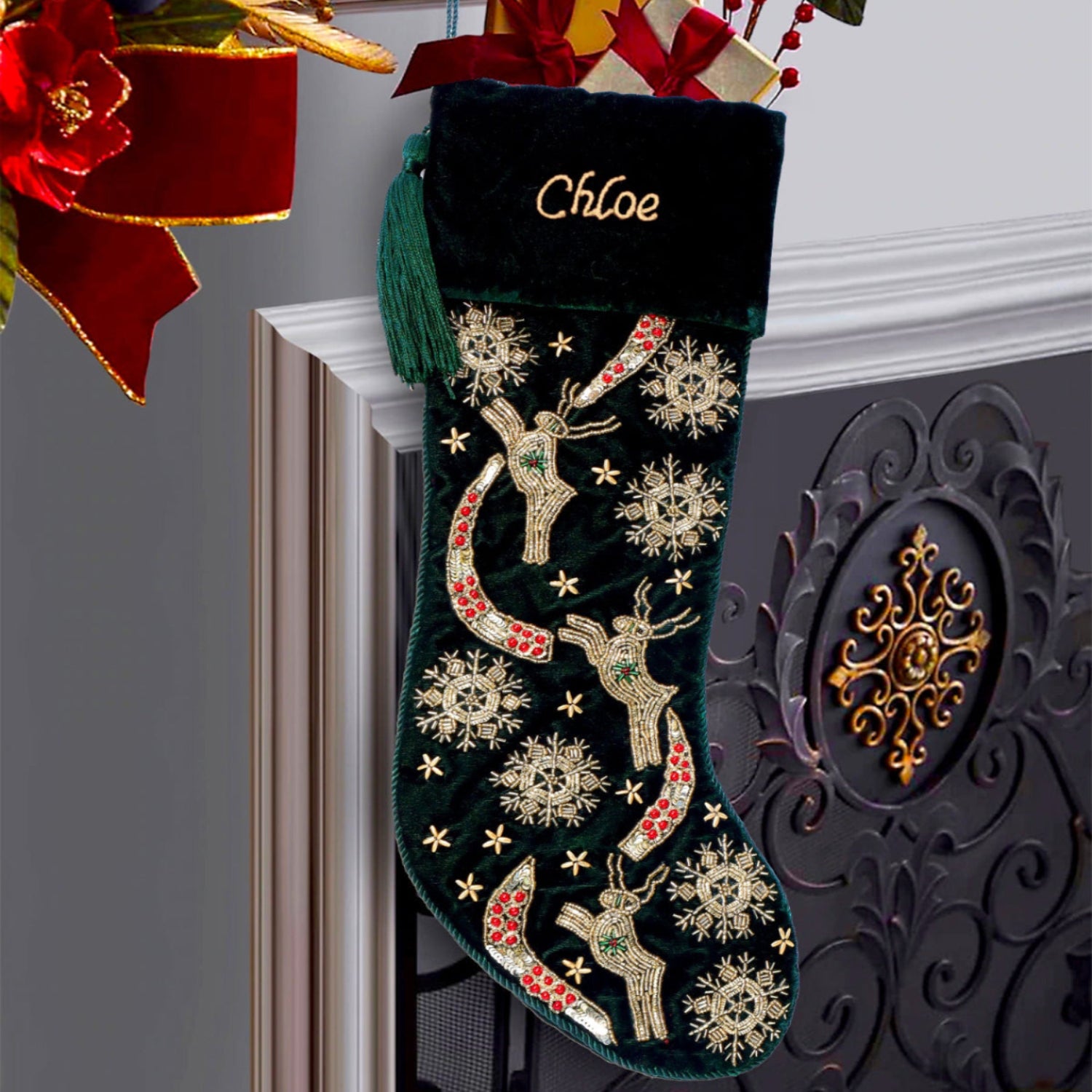 A dark green velvet Christmas stocking with embroidered gold and red reindeer, snowflakes, and stars, hanging on a fireplace mantel next to a black decorative screen. The stocking is personalized with the name 'Chloe' in gold embroidery at the top and is decorated with a gift box and ribbon.